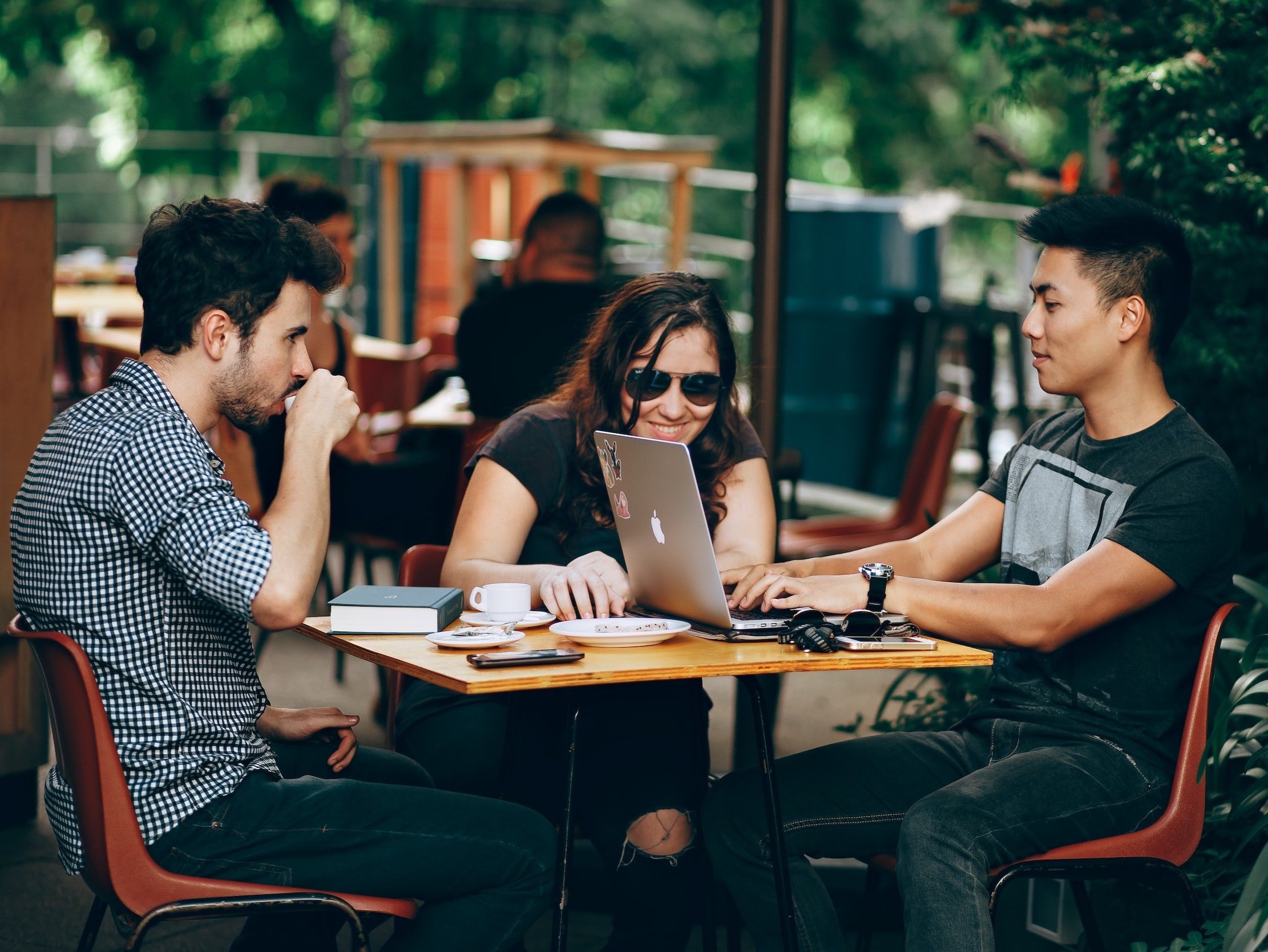 Three friends converse over coffee at a table while looking at an Apple Macbook.