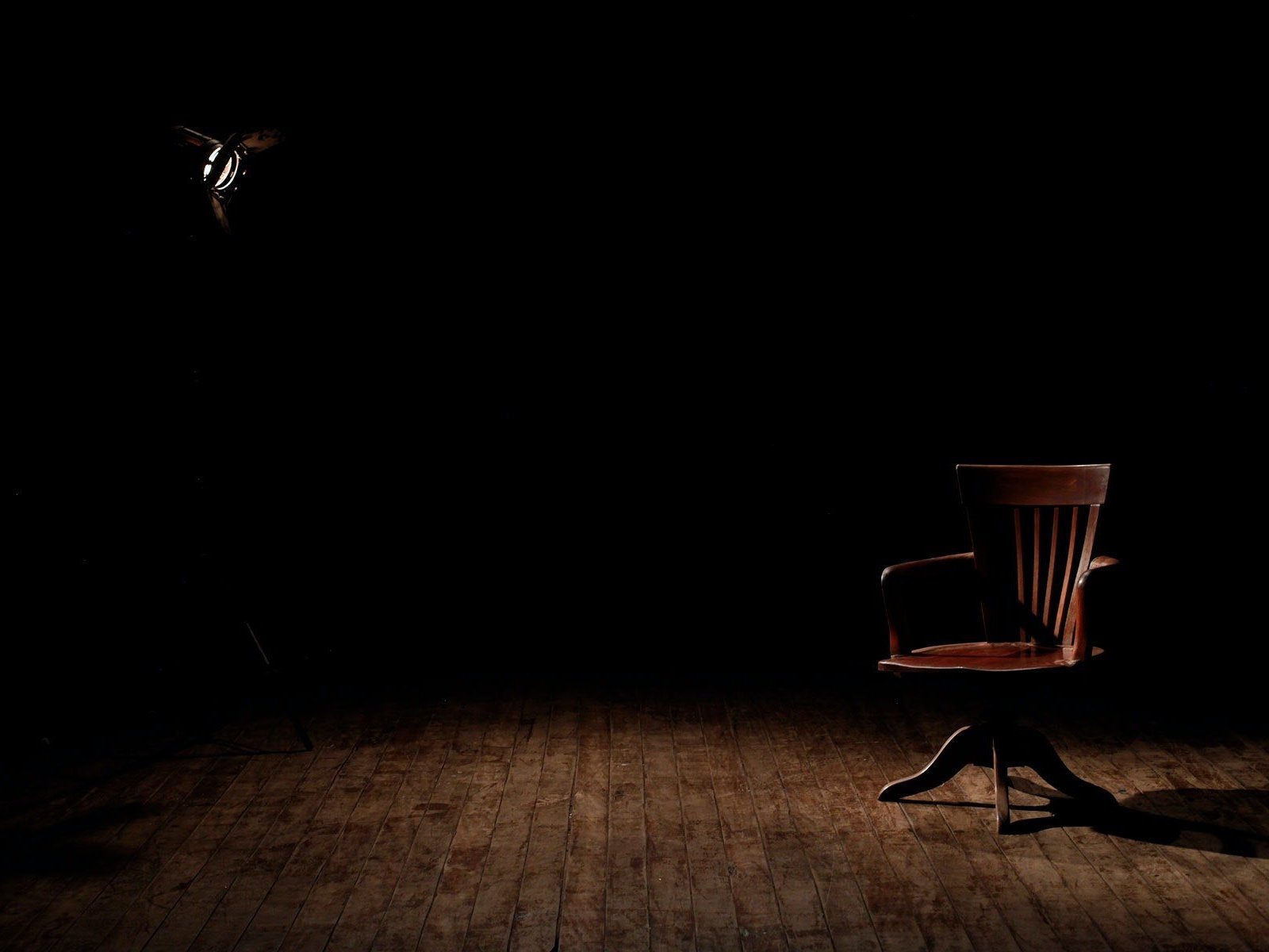 A spotlight illuminates an empty wooden armchair sitting on a brown wooden floor.