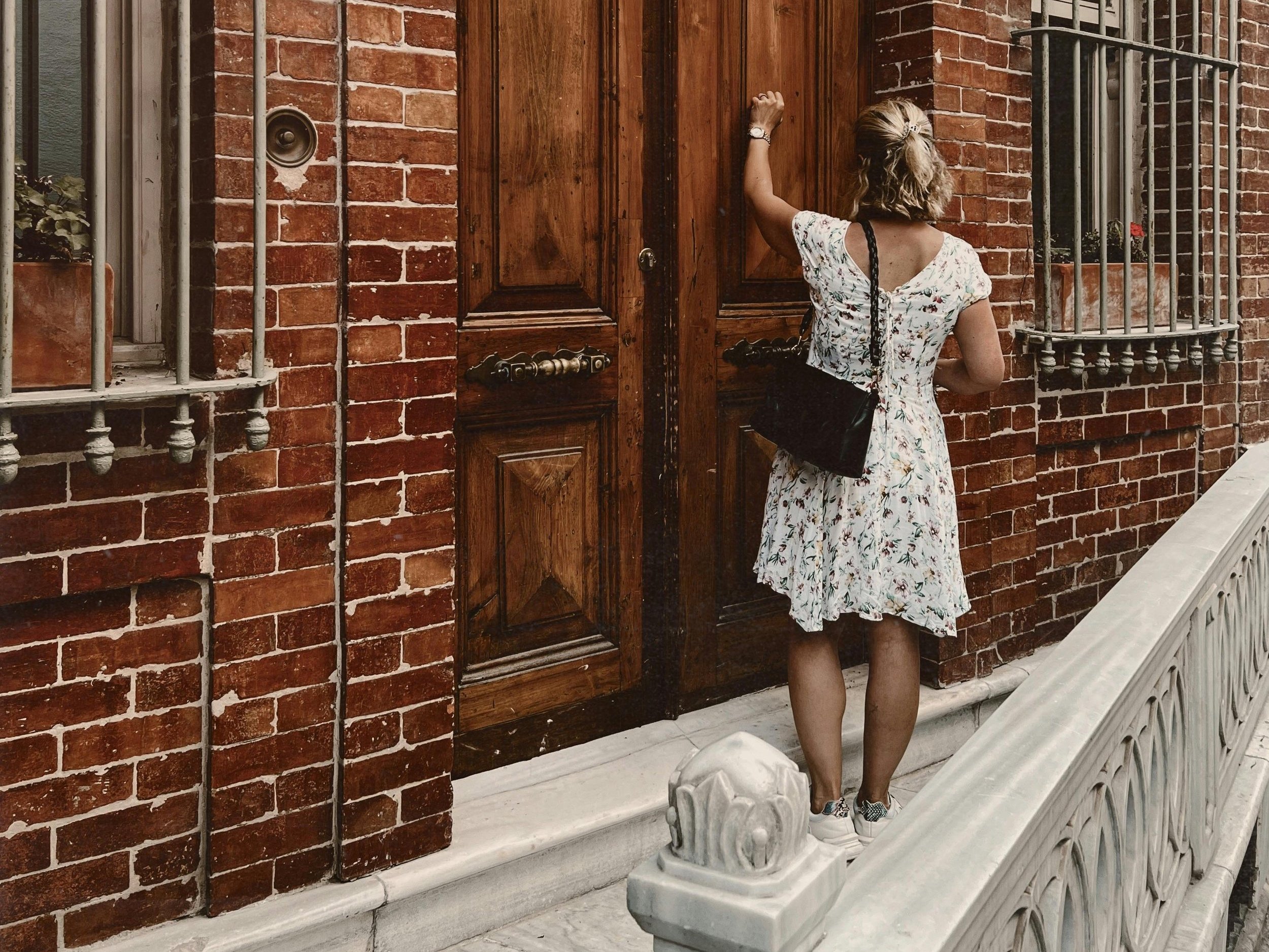 A woman in a floral dress knocking on a large wooden door of a brick building.