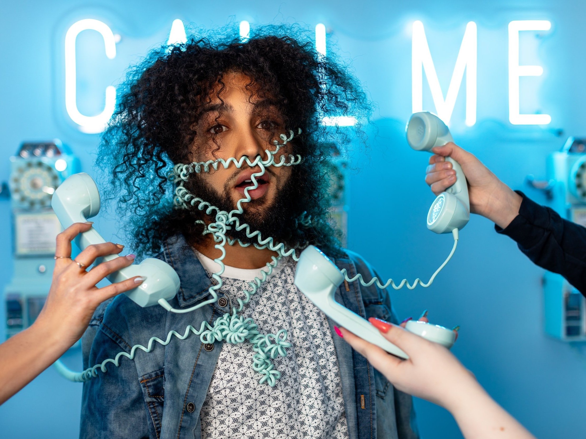 A man with multiple phone hand sets points to him, with telephone cables wrapped around him, and the word "Call Me" illuminated in neon lights up behind him.