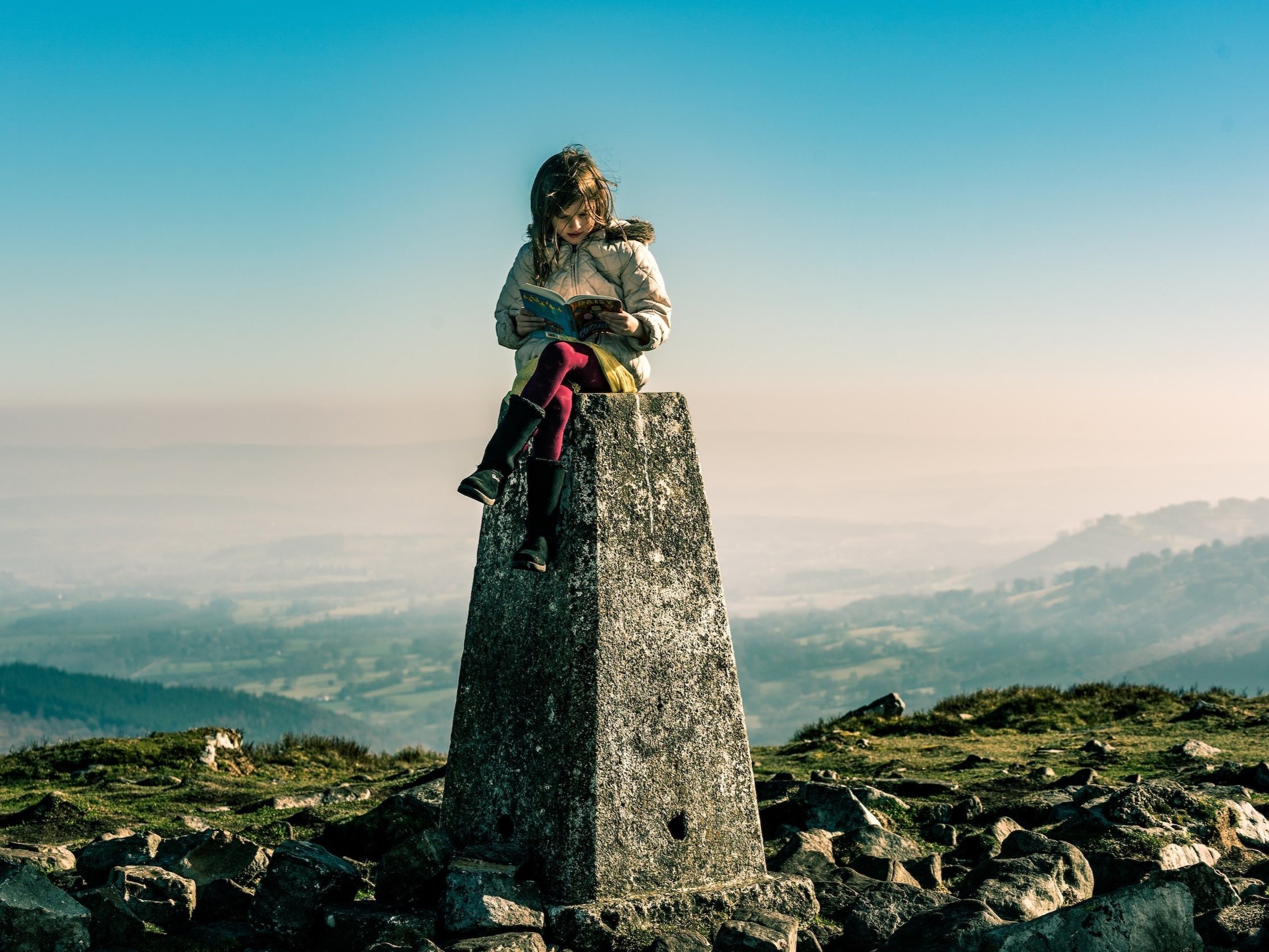 On a trig point, a young girl is reading a book.