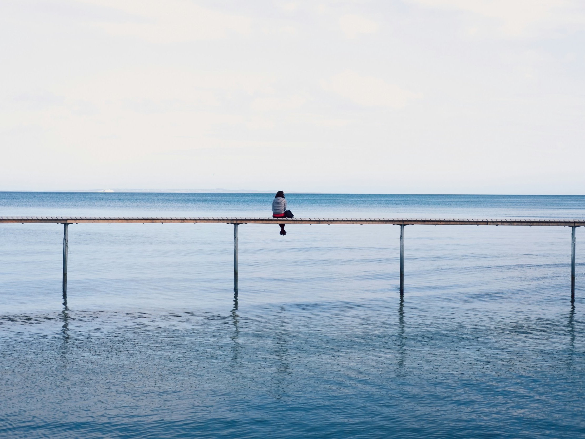 A person sits on a tranquil pier extending over calm blue waters under a clear sky, evoking a sense of solitude and reflection.