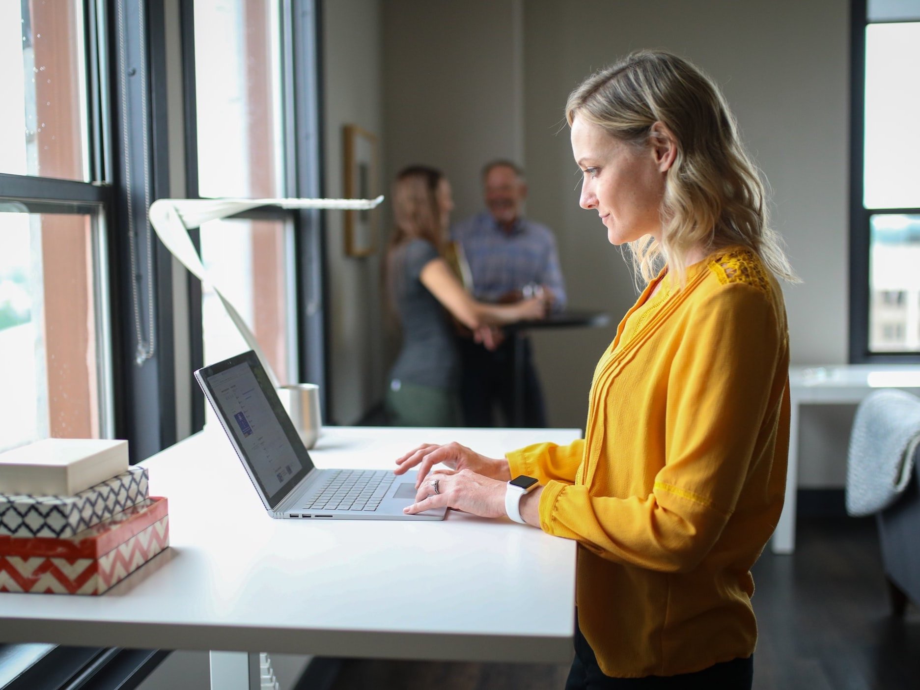 A woman works in the office while standing in front of a window at her standing desk.