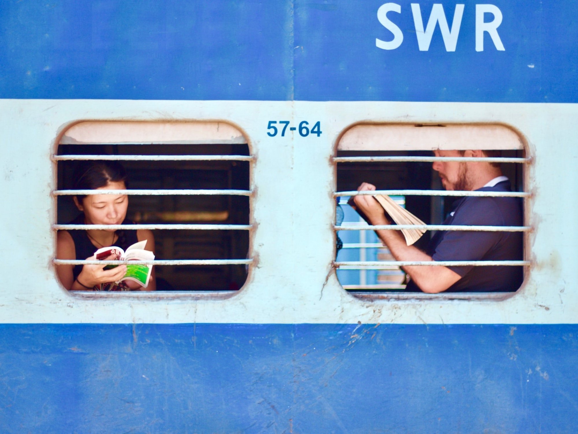 Two passengers are reading books in front of the windows of a train.