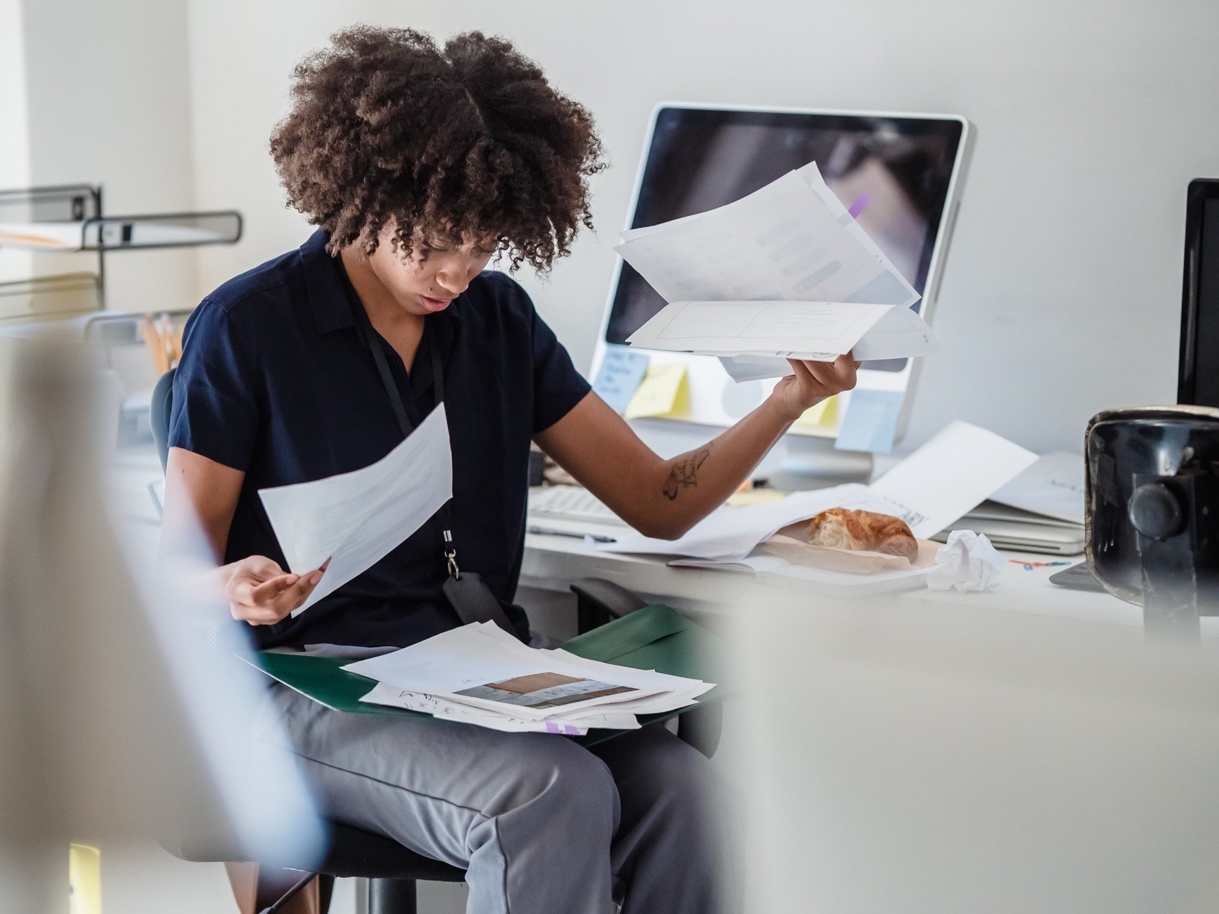 A woman in an office with a busy and cluttered desk is going through documents on her lap.