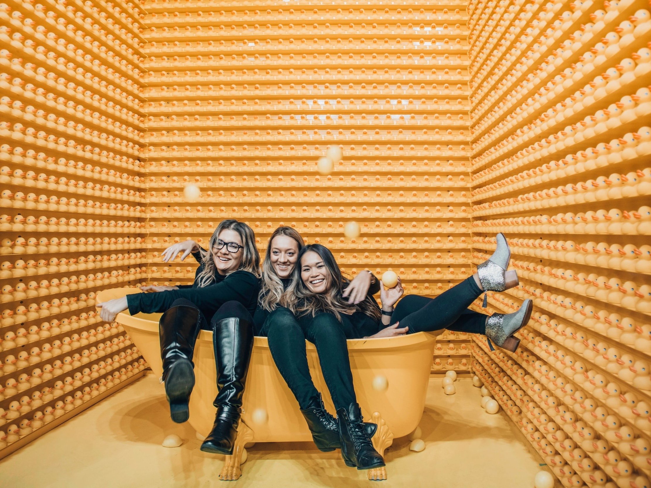 Three women are laughing while lying in an orange bath with their clothes on, rubber ducks lining the walls, and throwing plastic balls in the air.