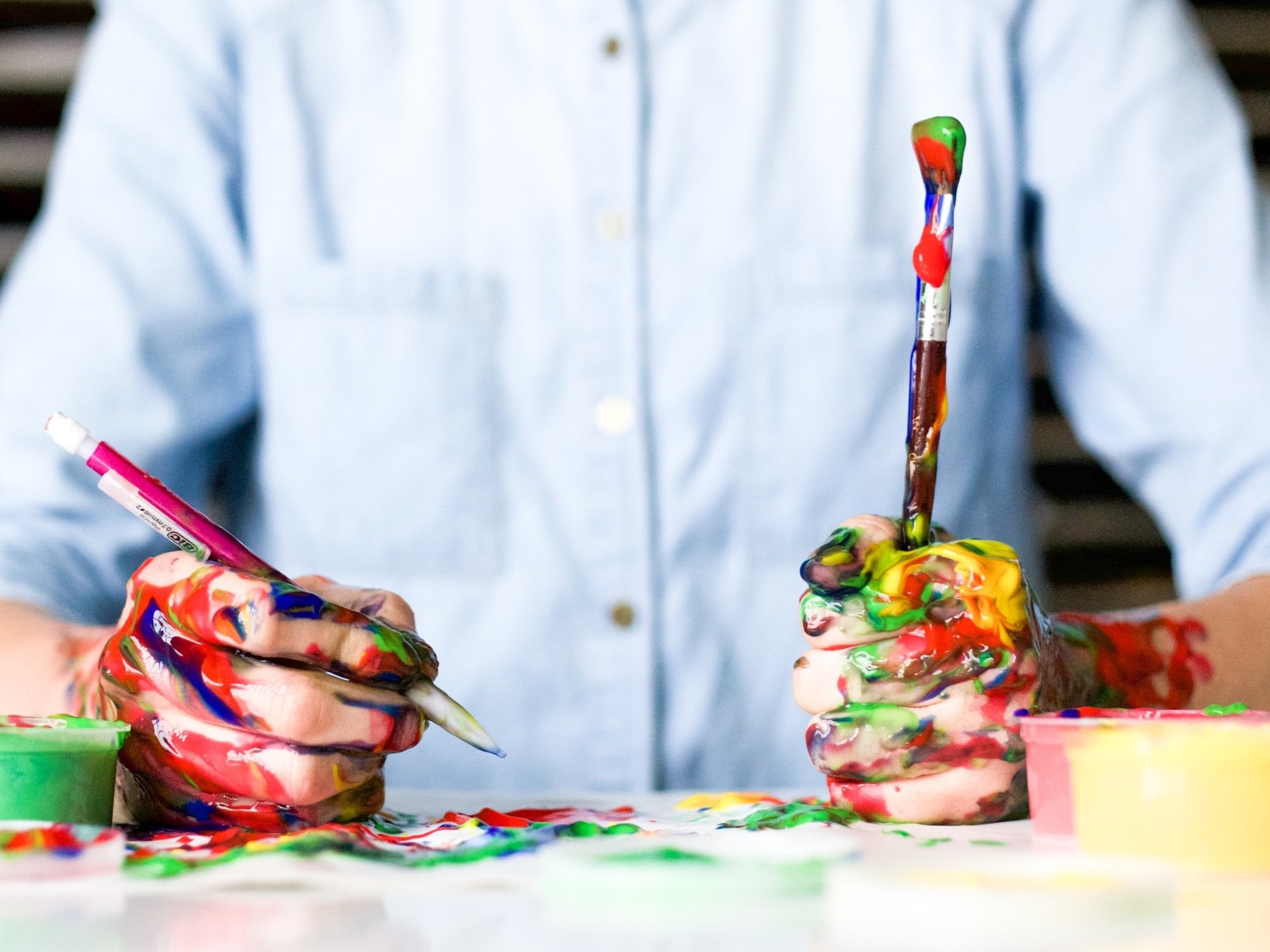 A person holding a paintbrush and a pencil, both of which are drenched in various paints.