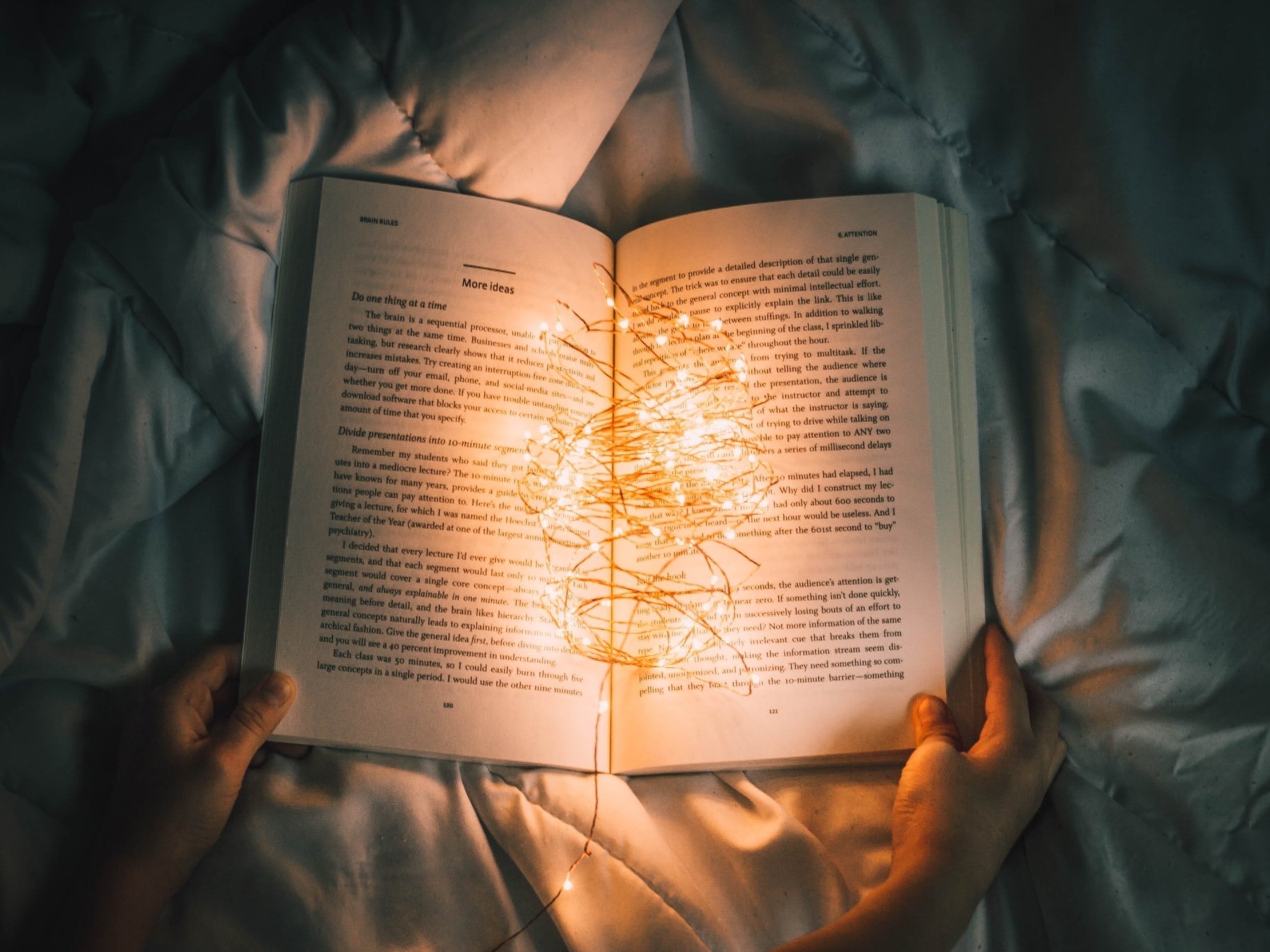 Someone reading a book in bed while a ball of glowing led lights illuminates the book.
