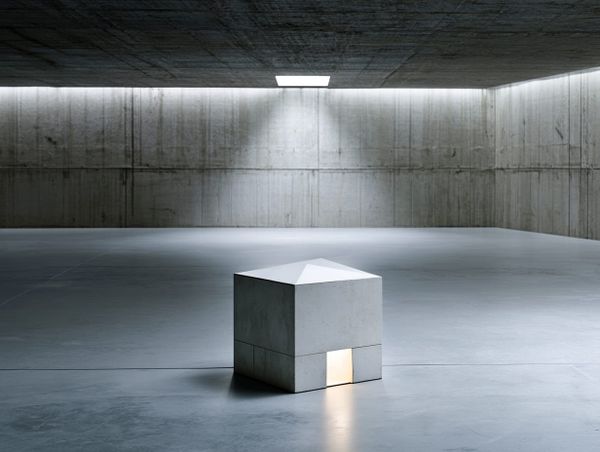 Small concrete structure with warm light glowing from a low doorway, dwarfed by a vast empty concrete hall.