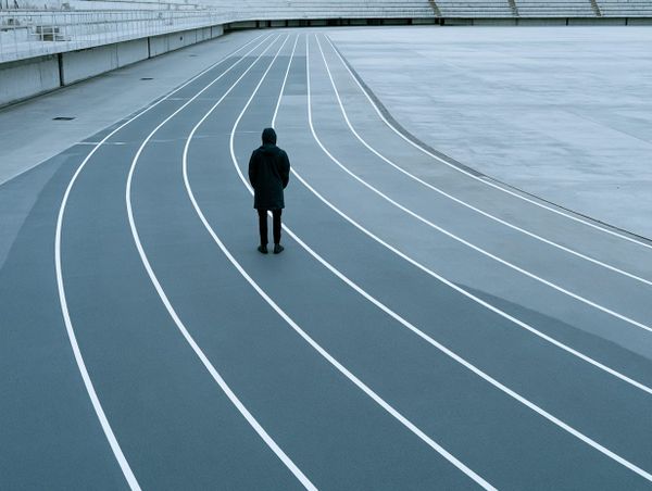 A figure stands still on an empty curved running track where the lanes bend and open into bare concrete.