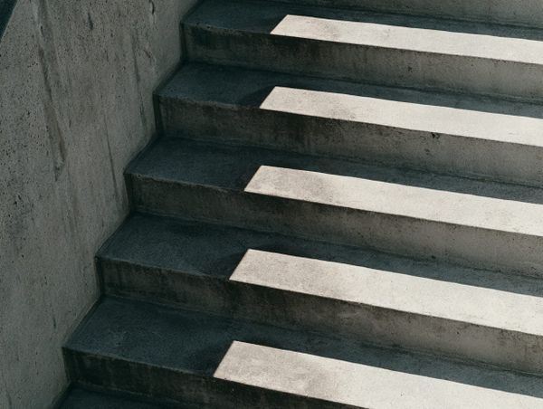 Concrete staircase in raking side light, pale slivers falling across each dark tread in a repeating rhythm of accumulated wear.