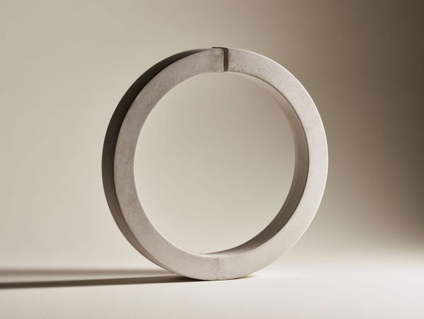 Pale sculptural ring standing upright with a clean vertical break in its form, soft window light on a neutral background.