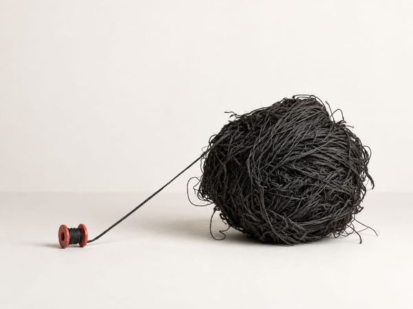 A small red wooden spool trails one black thread across a pale surface to a large tangled ball of black thread.