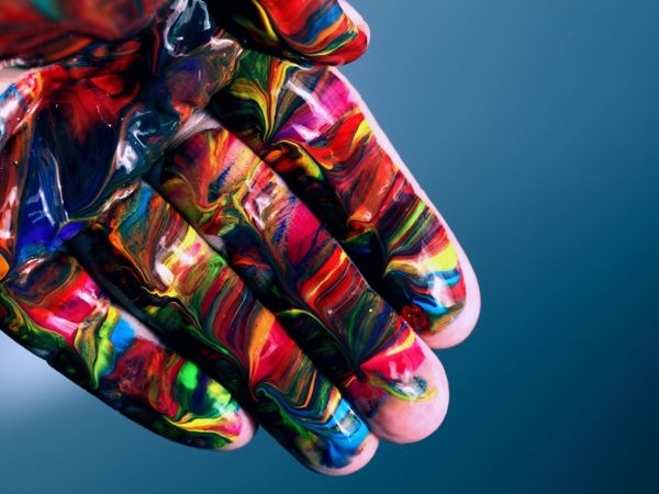 Open hand covered in vibrant, multi-colored paint - red, blue, yellow, green swirls and marbling on blurred blue background.