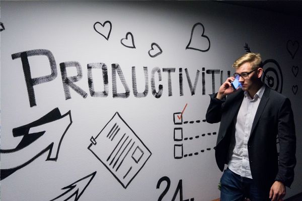 "Person in a suit on a phone call standing in front of a wall with the word 'PRODUCTIVITY' and various drawings and icons."