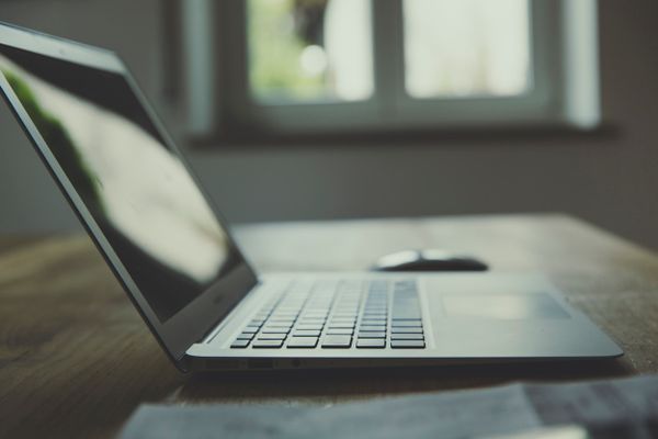 Laptop on a wooden desk with papers and a mouse, near a window with daylight.