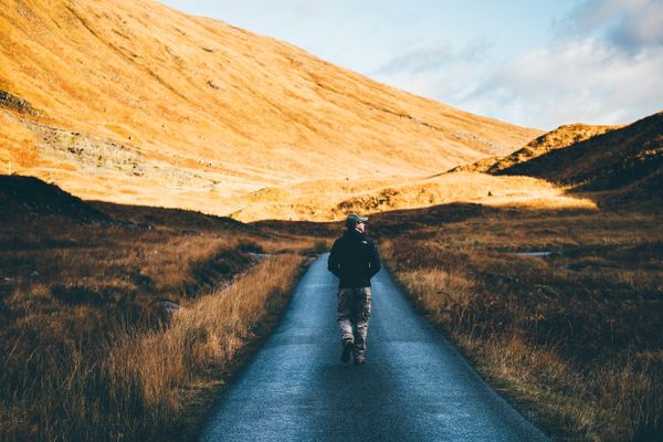 Man walking alone on a road in a scenic, sunlit mountainous landscape, symbolizing a break from digital life.