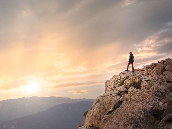 Silhouette of person standing on rocky cliff edge at sunset, overlooking mountains, symbolising seeking opportunities.