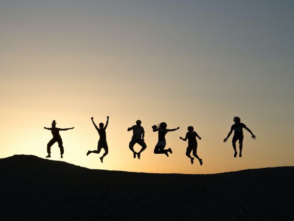 Silhouettes of six people jumping joyfully against an orange sky at sunset, conveying positivity and friendship.