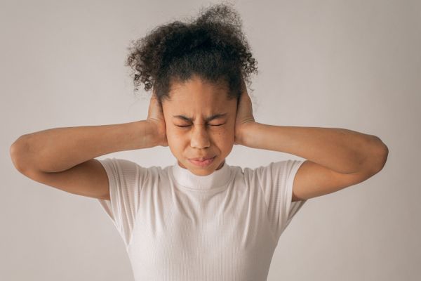 Young woman with curly hair covering her ears, eyes shut tight, wearing a white shirt, looking distressed.