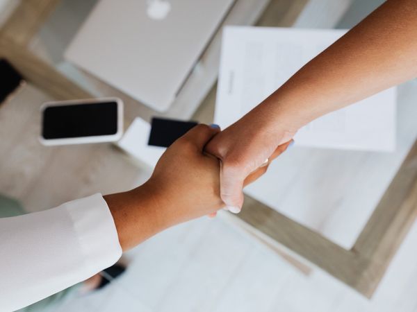 Close-up of two people shaking hands, with phones and paperwork on a table, suggesting a business meeting or agreement.