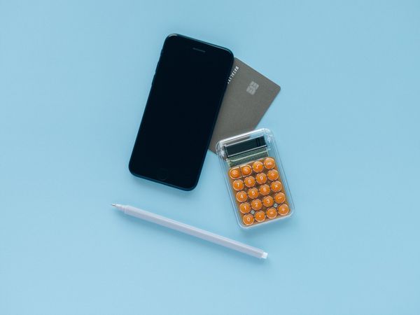 A smartphone, calculator, pen, and notebook on a light blue background, suggesting office work or financial calculations.