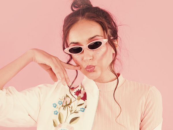 Young woman with red hair and white sunglasses, her face adorned with pearls, poses playfully on a pink background.