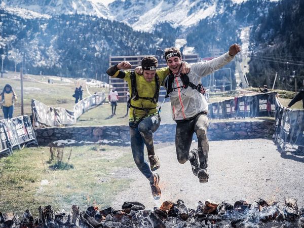 Outdoor enthusiasts leap over obstacles at snowy mountain event, rocky terrain in background.