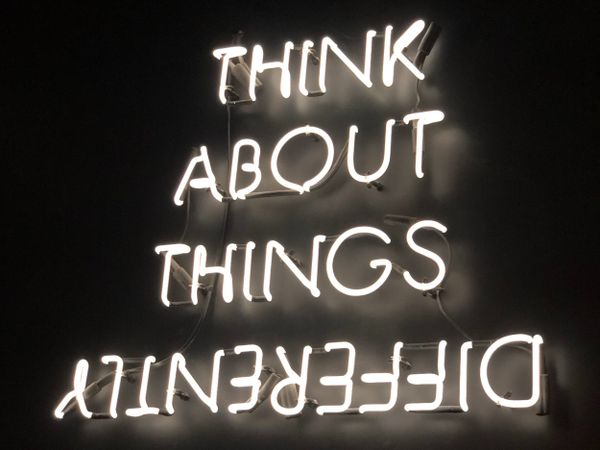 Neon sign: "Think about things differently", implying novel perspectives, not just more effort, drive breakthroughs.