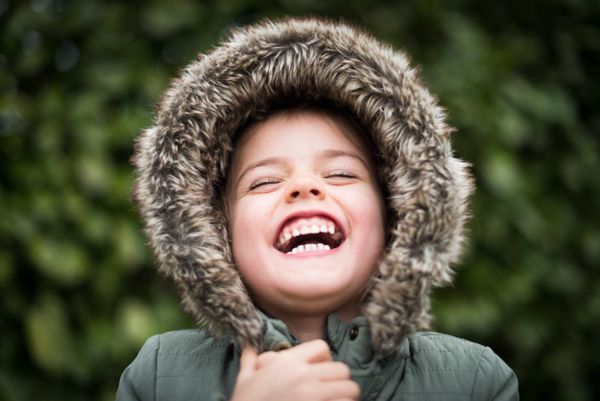 Smiling child in fur-lined hooded coat laughs joyfully outside surrounded by green foliage.