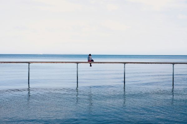 Person sits on serene pier over calm blue water under clear sky, conveying solitude and reflection.