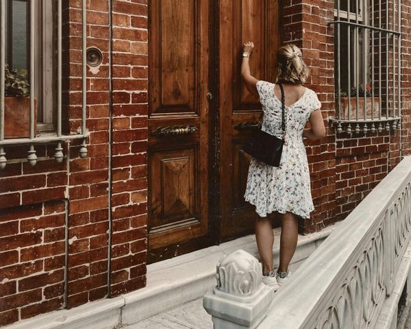 A woman in a floral dress knocking on a large wooden door of a brick building.