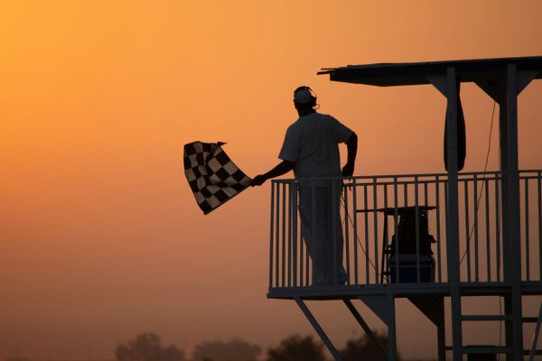Silhouetted race official waves checkered flag on platform at sunset, signaling start or end of auto racing event.