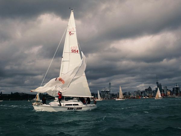 Sailboat with crew navigates choppy waters against cloudy sky, city skyline visible in the background.