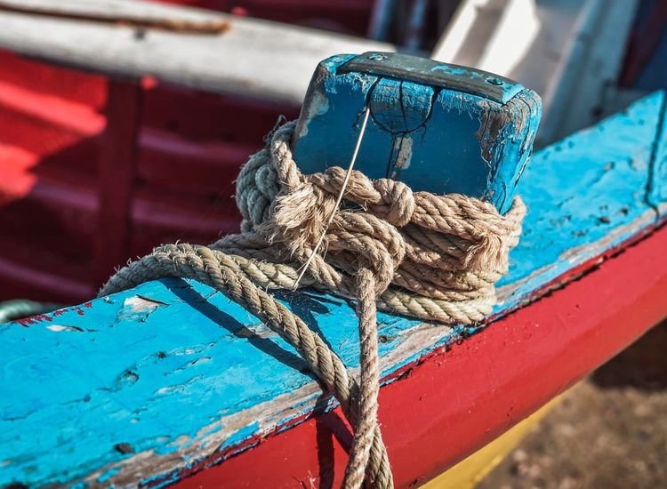 A wooden fishing boat on the shore with worn out blue and red paint, rope tied to the boat