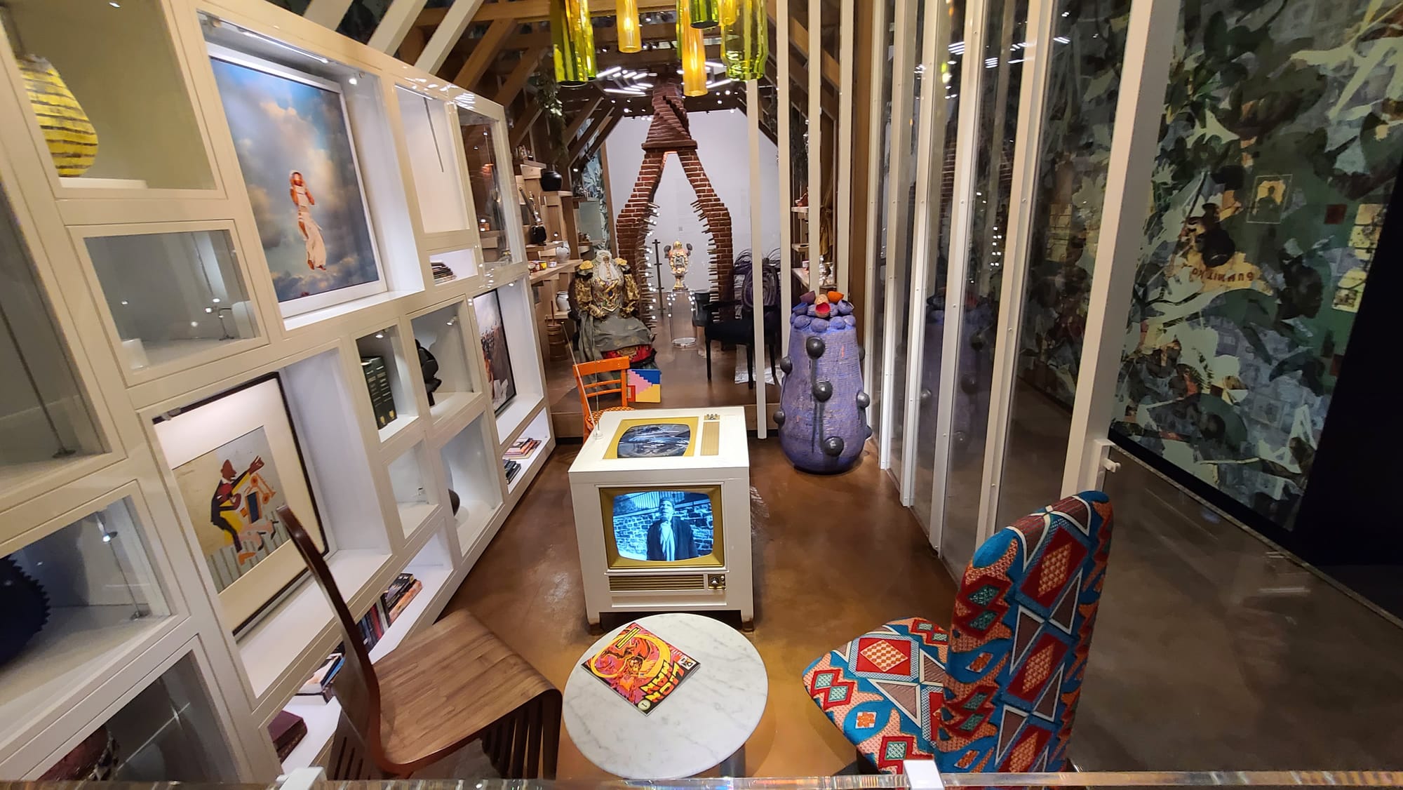 A  museum exhibition with white shelving and framed art on the left side, two chairs on the floor, an old-fashioned television in the middle of the room. A purple sculpture in the back underneath a chandelier made of green and yellow glass, and a brick open sculptural chimney in the background.