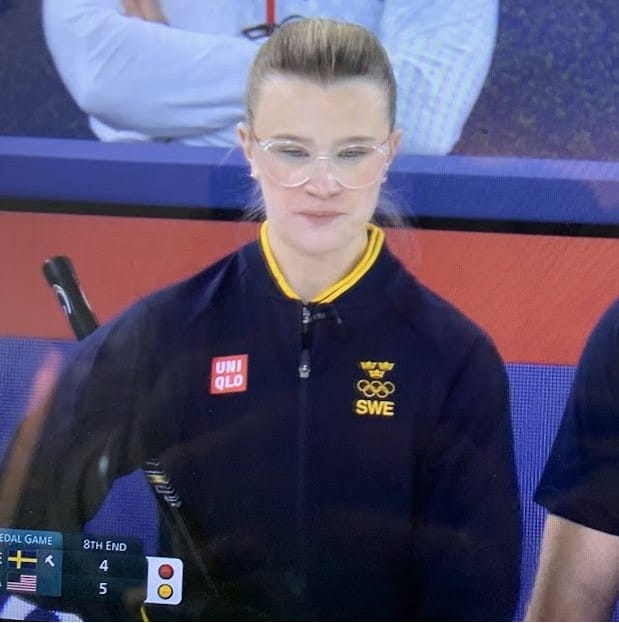 Image of a Swedish curler on the women's Winter Olympics team looking intently at the ice behind a cute pair of large, clear glasses.