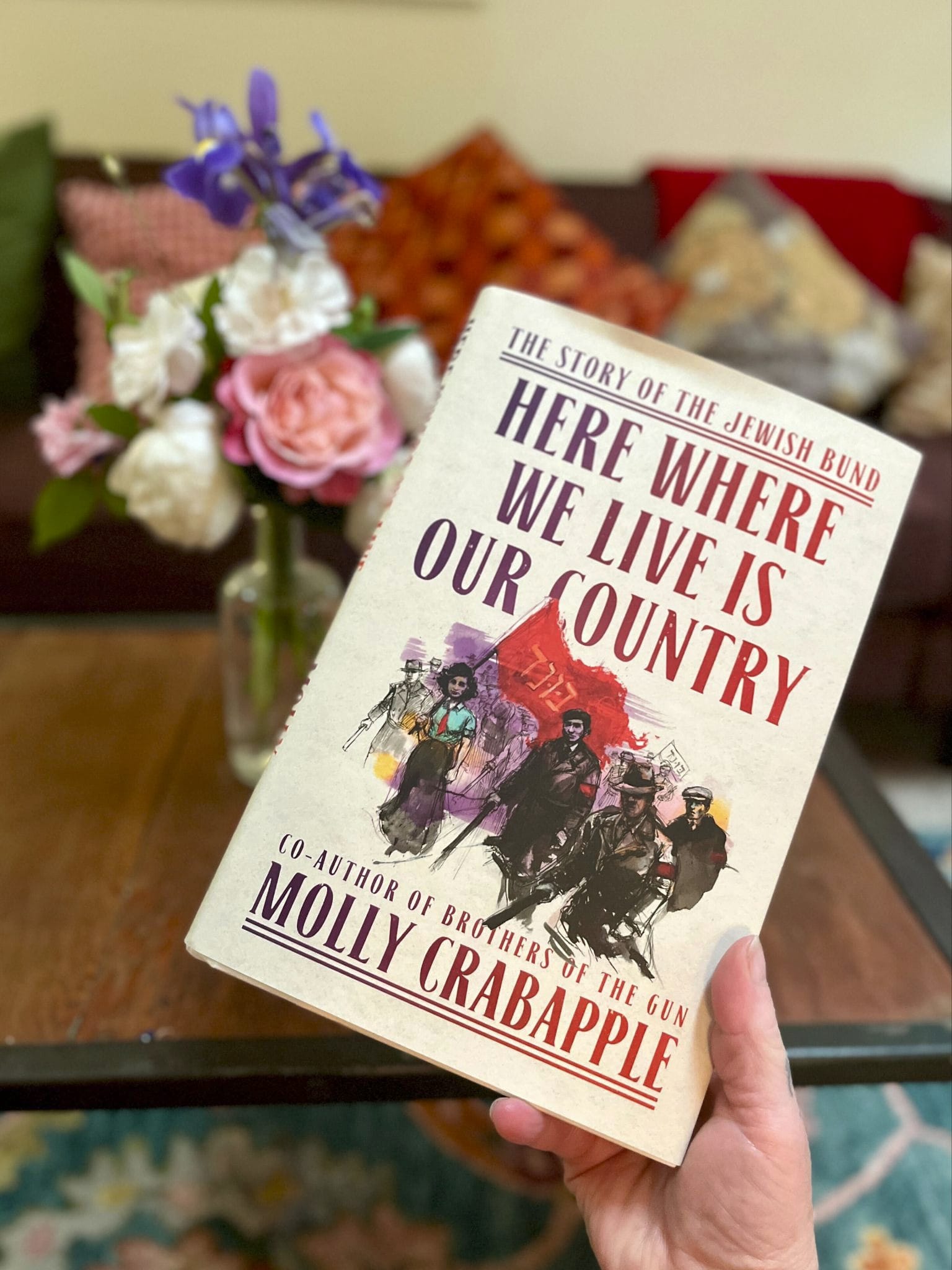 foreground, a hand holds a white book with red and purple text over a watercolor image of people in military uniform.