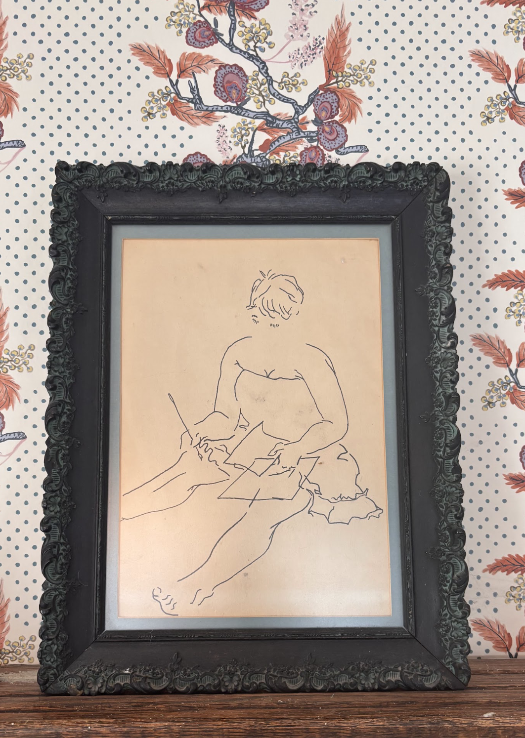 A line drawing on parchment inside an embellished black frame. In it, a woman sits with her legs extended looks down at the papers on her lap. The lower half of her face remains unknown.
