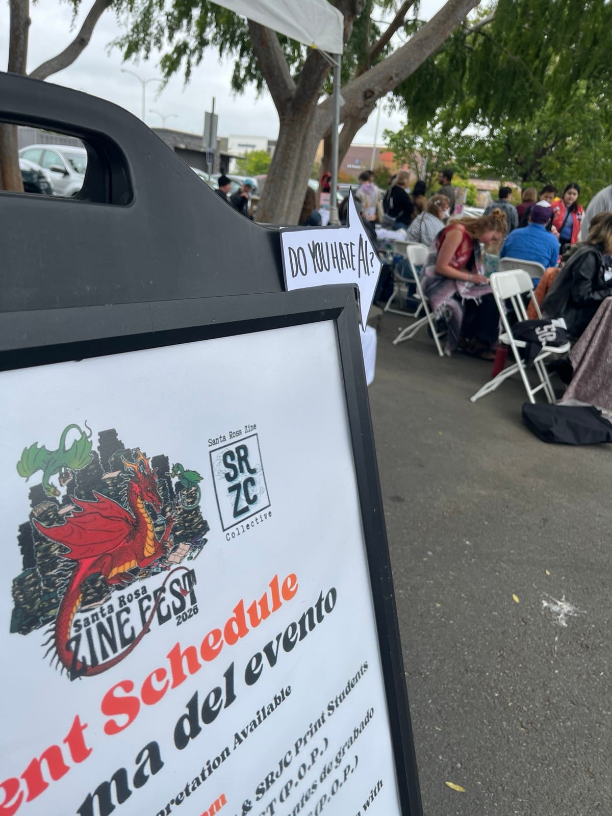 A sign for the Santa Rosa Zine fest is black and white and red, with an image of a dragon reading zines, and an arrow at the top says "Do you hate AI?" pointing toward a parking lot filled with people at chairs and tables.