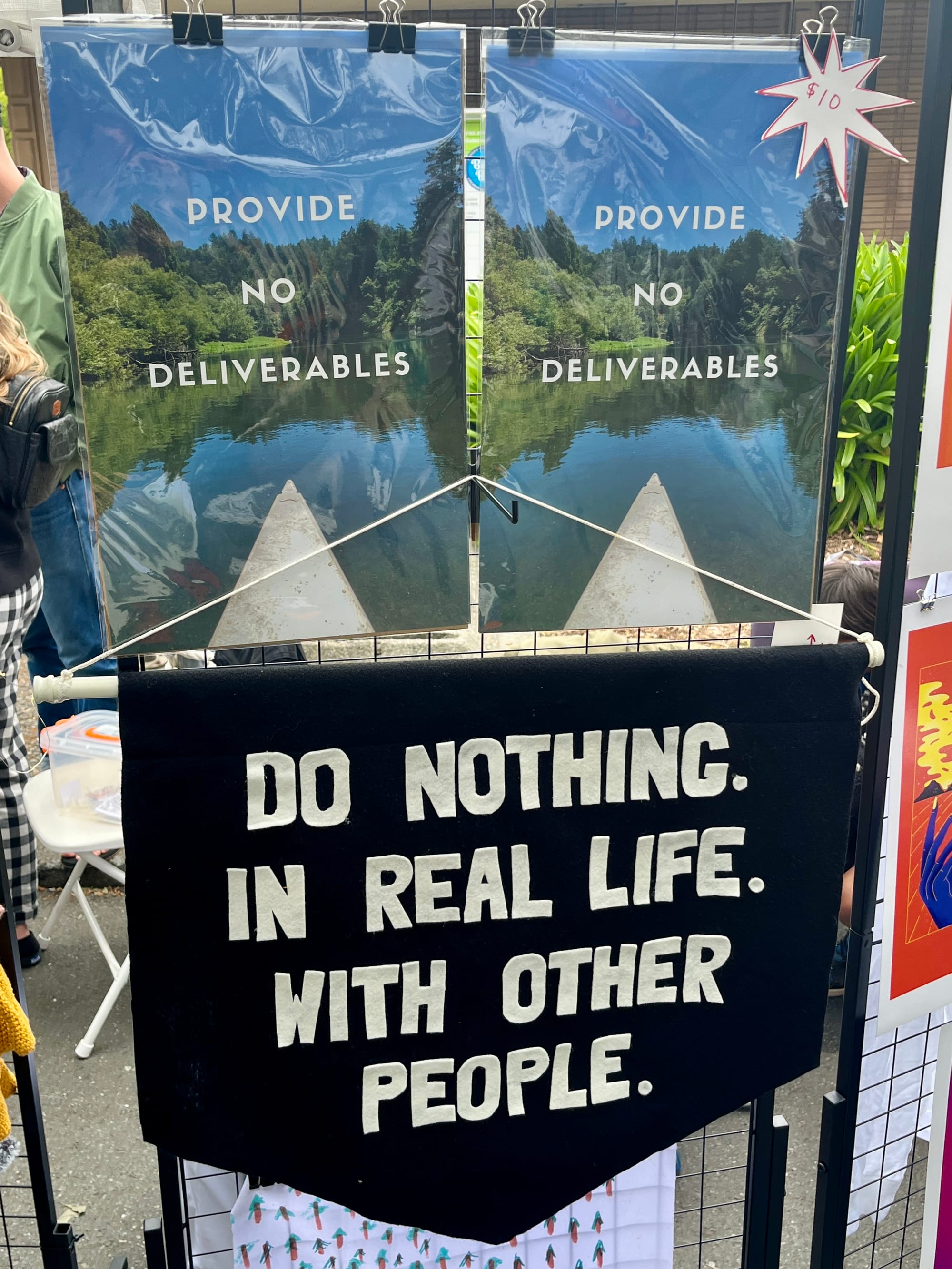 A display of art on black wire stands. Two posters with a nature scene over which text says "provide no deliverables" hang over a black flag hanging that reads: "Do Nothing. In Real Life. With Other People." Other art and crocheted cute animals hang nearby.