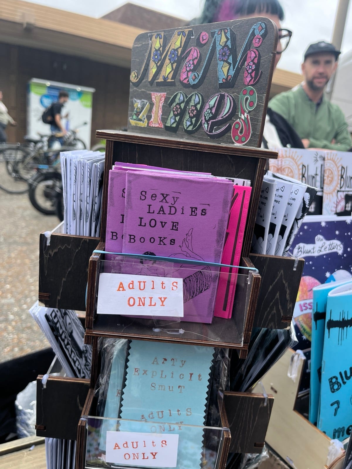 A tower says Mini Zines in collage cutout letters at the top, with two "Adults Only" shelves with zines, one is purple and says Sexy Ladies Love Books with an outline of a naked woman. The other is blue and says Arty Explicit Smut. In the background are more shelves holding more zines.