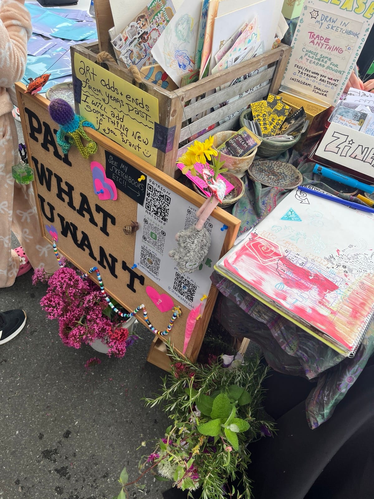 A very crowded table, with buckets of plants and flowers on the concrete, a bulletin board on teh front that reads PAY WHAT U WANT in black cutout letters, and baskets and piles of colorful zines on top.