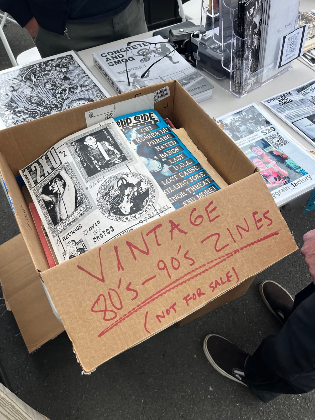 A cardboard box has red marker lettering spelling out: VINTAGE 80's - 90's ZINES (NOT FOR SALE), inside are piles of old photocopied zines, many with photographs of people playing guitar.