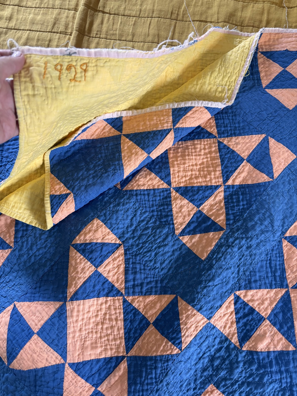 A beautiful blue quilt with peach-colored triangles distributed throughout it. In the top right corner, a person displaying the quilt's underside shows the number '1929.'