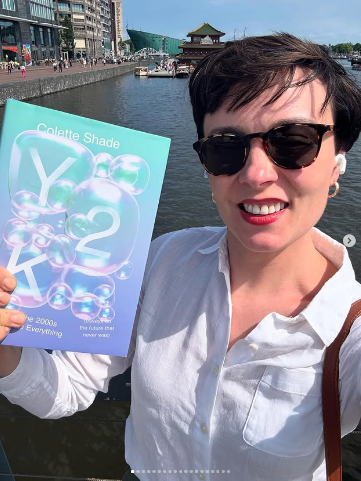 A white woman with short dark hair and sunglasses holds a book up while standing by a river.