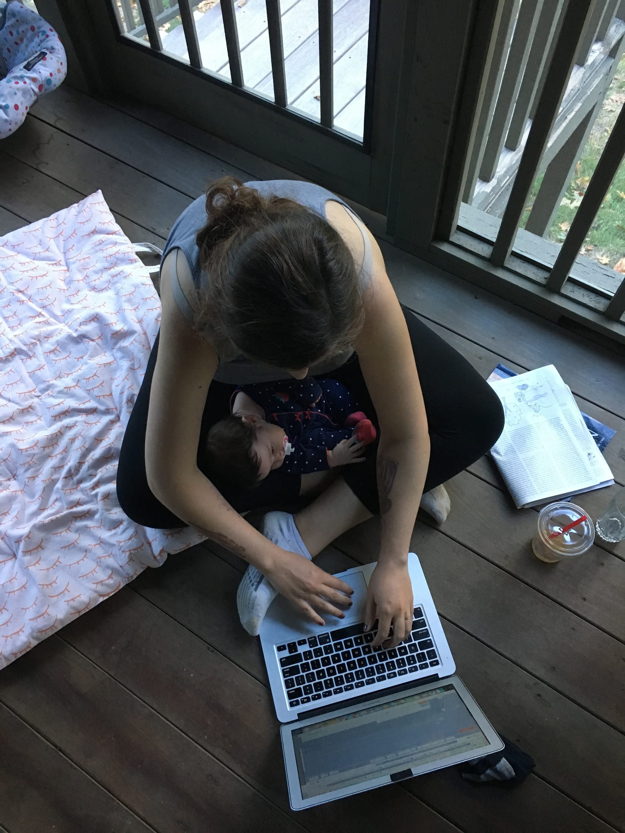 Overhead shot of a woman looking down on her laptop while she writes on a wooden deck. A baby is nestled between her crossed legs.