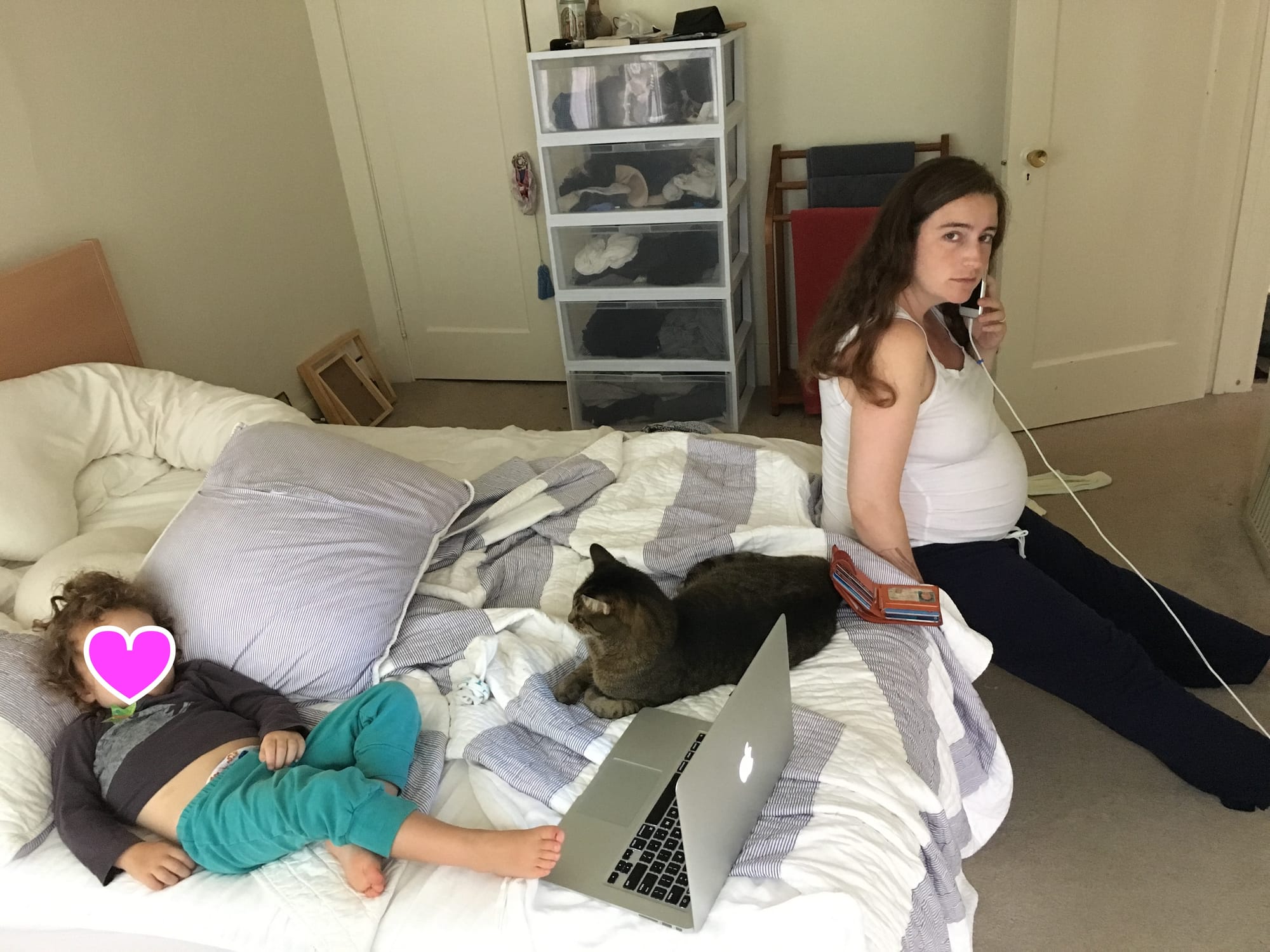 A photo of a pregnant white woman seated at the edge of her bed. She's looking directly at the camera with a smartphone in her hand and her wallet open beside her. Also, there is a cat.