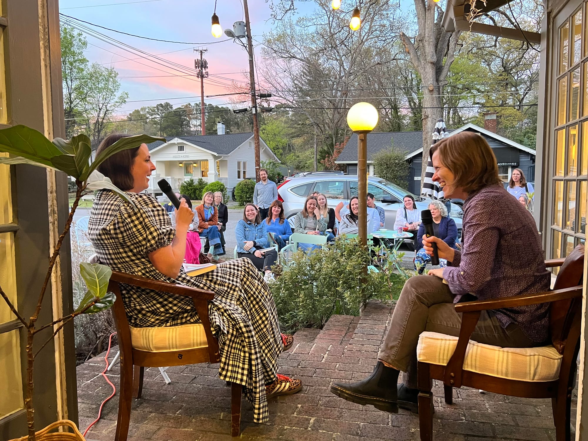 Two women sit in wooden armchairs on a rustic brick patio at dusk facing each other while holding microphones. In the background, a seated audience watches the conversation.