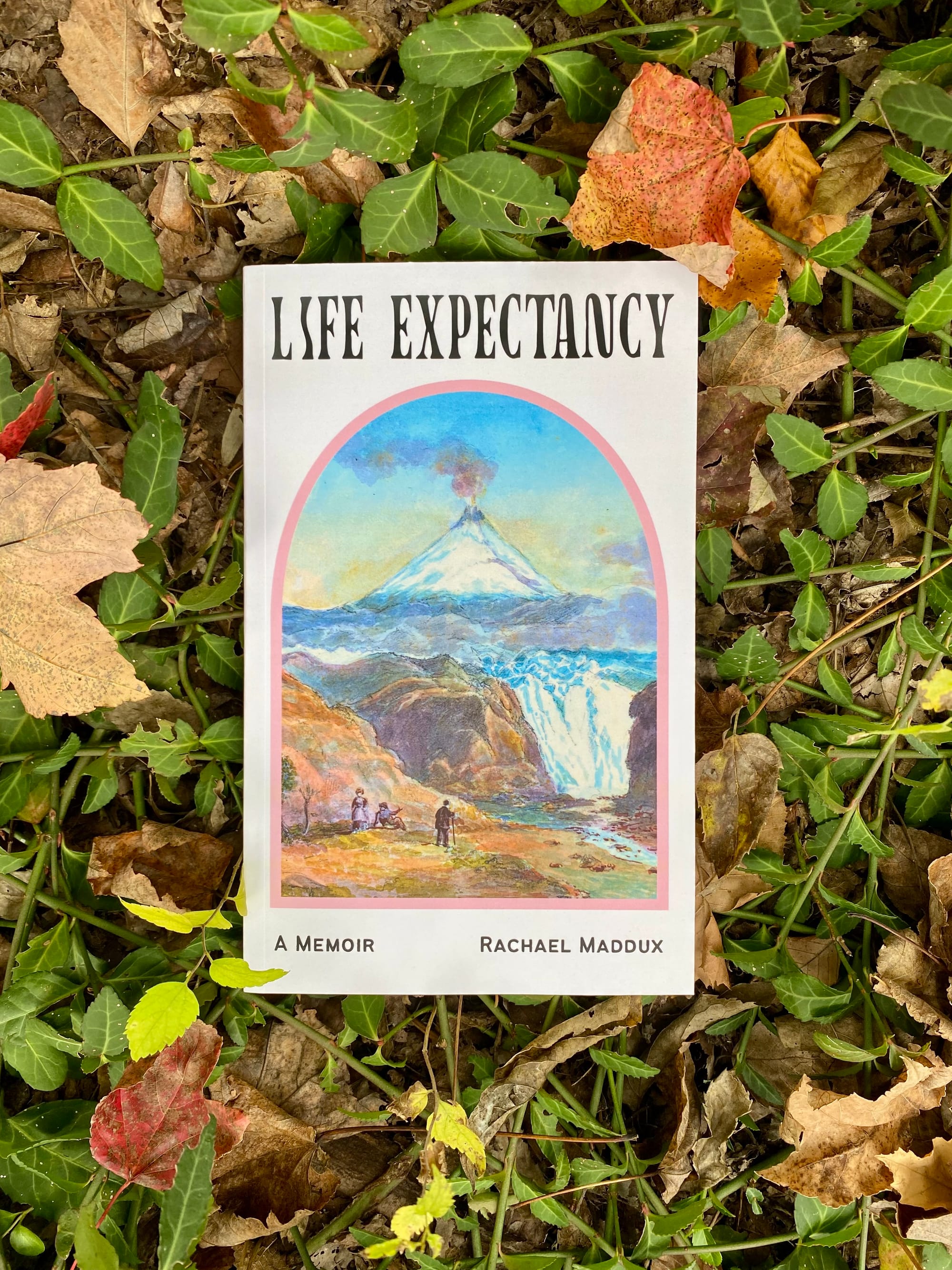 A book lies on a bed of autumn leaves mixed with  greenery. "LIFE EXPECTANCY" is written in black, tall letters. "A MEMOIR" and "RACHAEL MADDUX" are printed in smaller type at the bottom. 